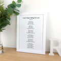 May include: A white framed print featuring the lyrics to "Can't Help Falling In Love" by Elvis Presley. The print is displayed on a wooden surface, with a decorative letter and greenery in the background. The text is in black.