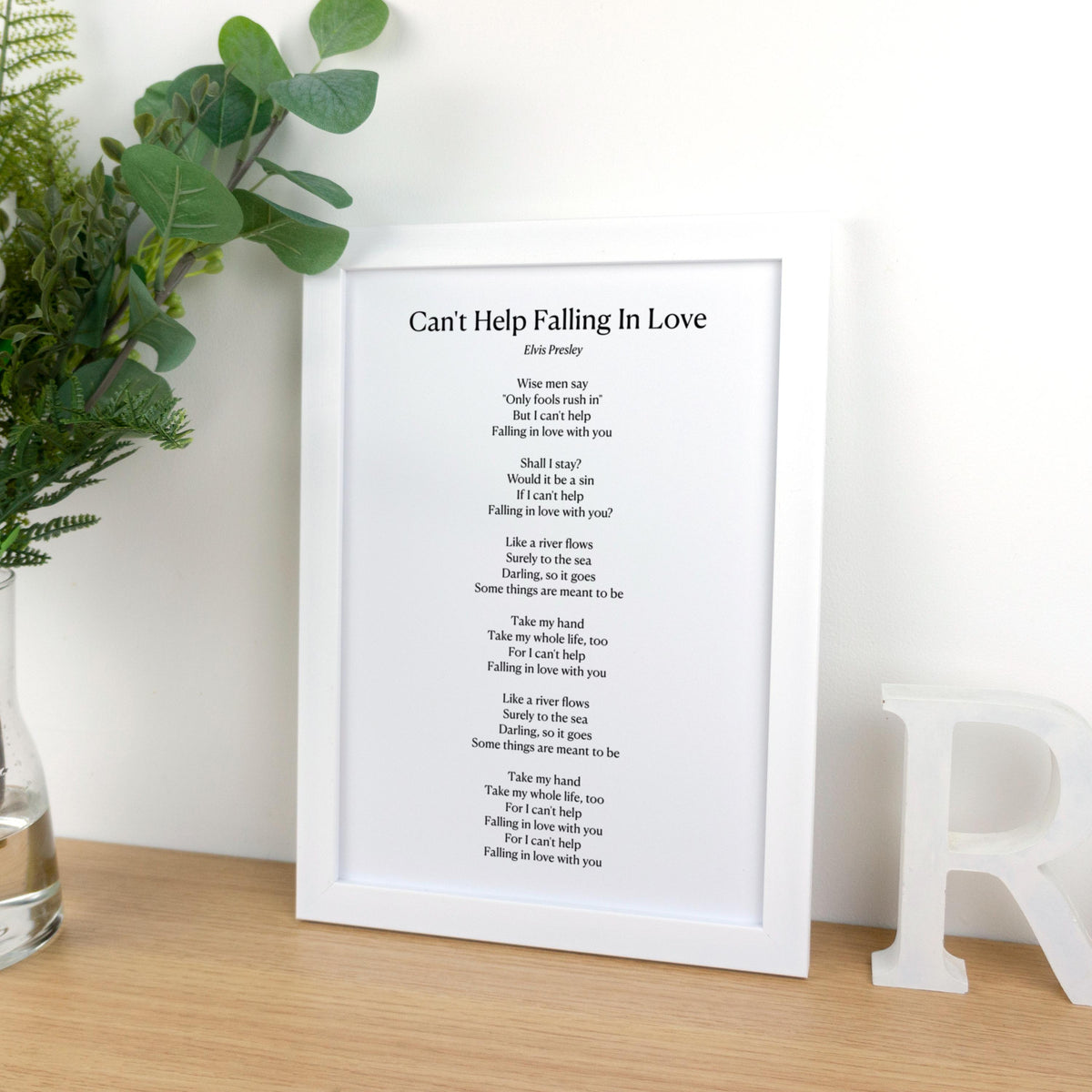 May include: A white framed print featuring the lyrics to "Can't Help Falling In Love" by Elvis Presley. The print is displayed on a wooden surface, with a decorative letter and greenery in the background. The text is in black.